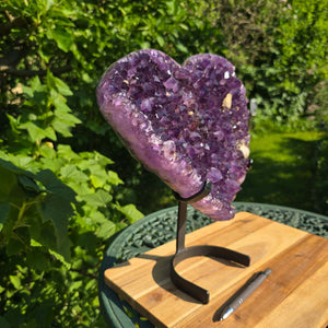 Amethyst Druse Amethyst Geode in Herzform mit Metallsockel 33x29x13cm - NR-17 auf einem stabilen Halter, umgeben von Natur.