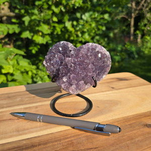 Amethyst Druse Amethyst Geode in Herzform mit Metallsockel 11x10x9cm - NR-48 auf einem Holztisch mit Stift.