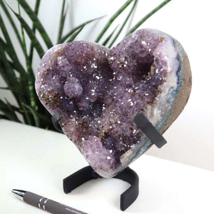 Amethyst Druse Amethyst Geode in Herzform mit Metallsockel 14x12cm - NR-10 auf schwarzem Ständer mit Pflanzen im Hintergrund