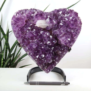 Amethyst Druse Amethyst Geode in Herzform mit Metallsockel 21x20cm - NR-25, auf schwarzem Ständer mit Pflanzen im Hintergrund.