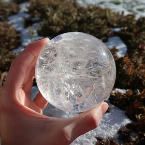 Bergkristall Kugel – Natürliche Kristallkugel 75mm Durchmesser Nr. 31 in einer Hand im Freien mit schnee- und pflanzenbedecktem Hintergrund