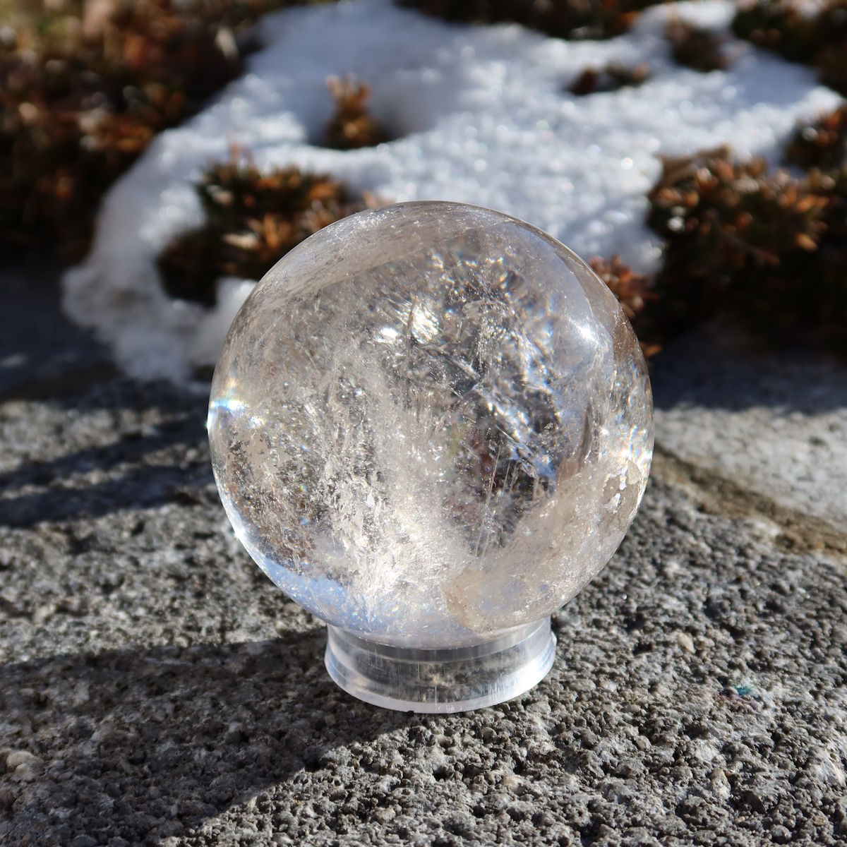Bergkristall Kugel – Natürliche Kristallkugel 60mm Durchmesser Nr. 41, klare Kristallkugel auf Steinuntergrund mit Schnee im Hintergrund.