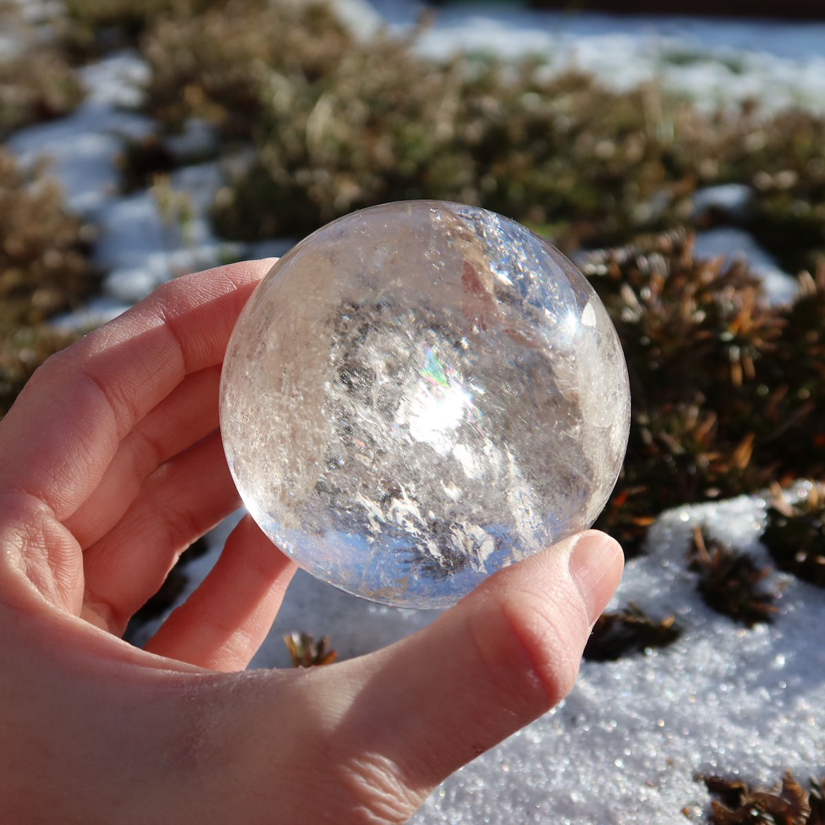 Bergkristall Kugel – Natürliche Kristallkugel 60mm Durchmesser Nr. 41, klare Kristallkugel auf Steinuntergrund mit Schnee im Hintergrund.