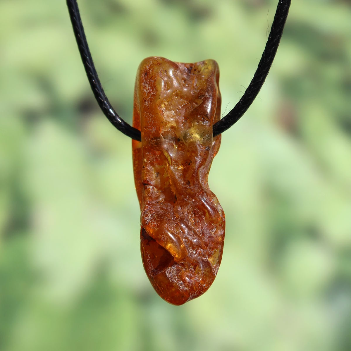 Natural amber pendant 46mm on black cord with unique texture, highlighting warmth and confidence, set against a blurred green background.