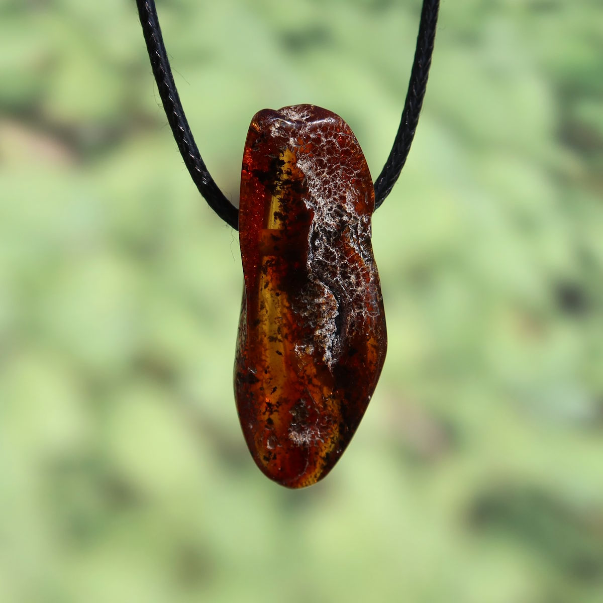 Natürlicher Bernstein Anhänger Unikat Nr. 20, 45mm, hervorgehoben auf dunklem Seil, transparente Textur im grünen Hintergrund
