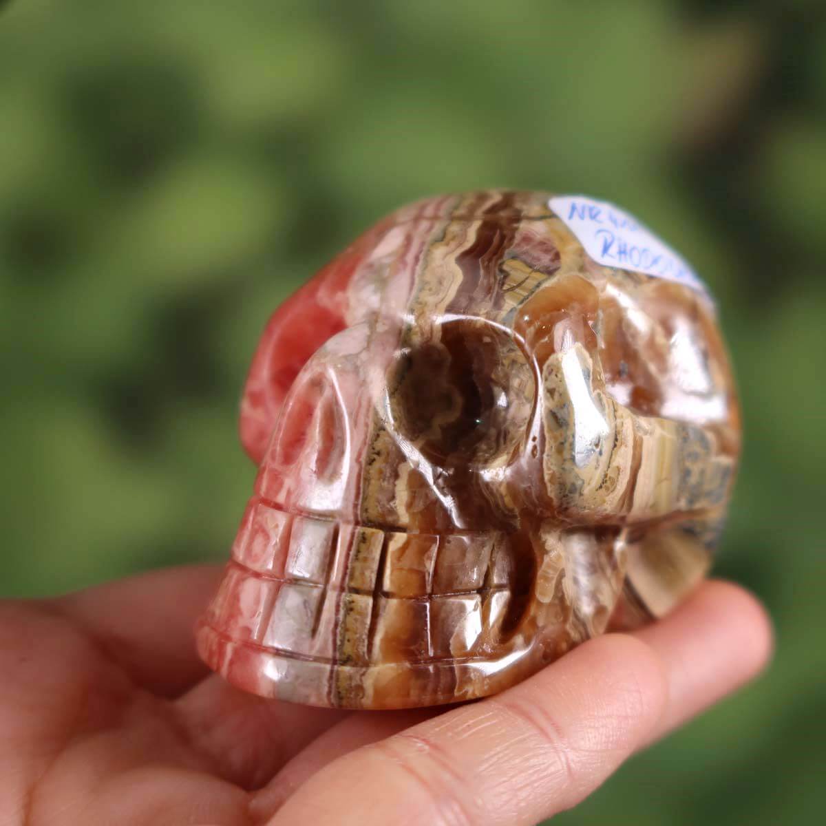 Handcrafted rhodochrosite crystal skull with unique shape, collector's item, 5x4 cm, held in hand against a green background.