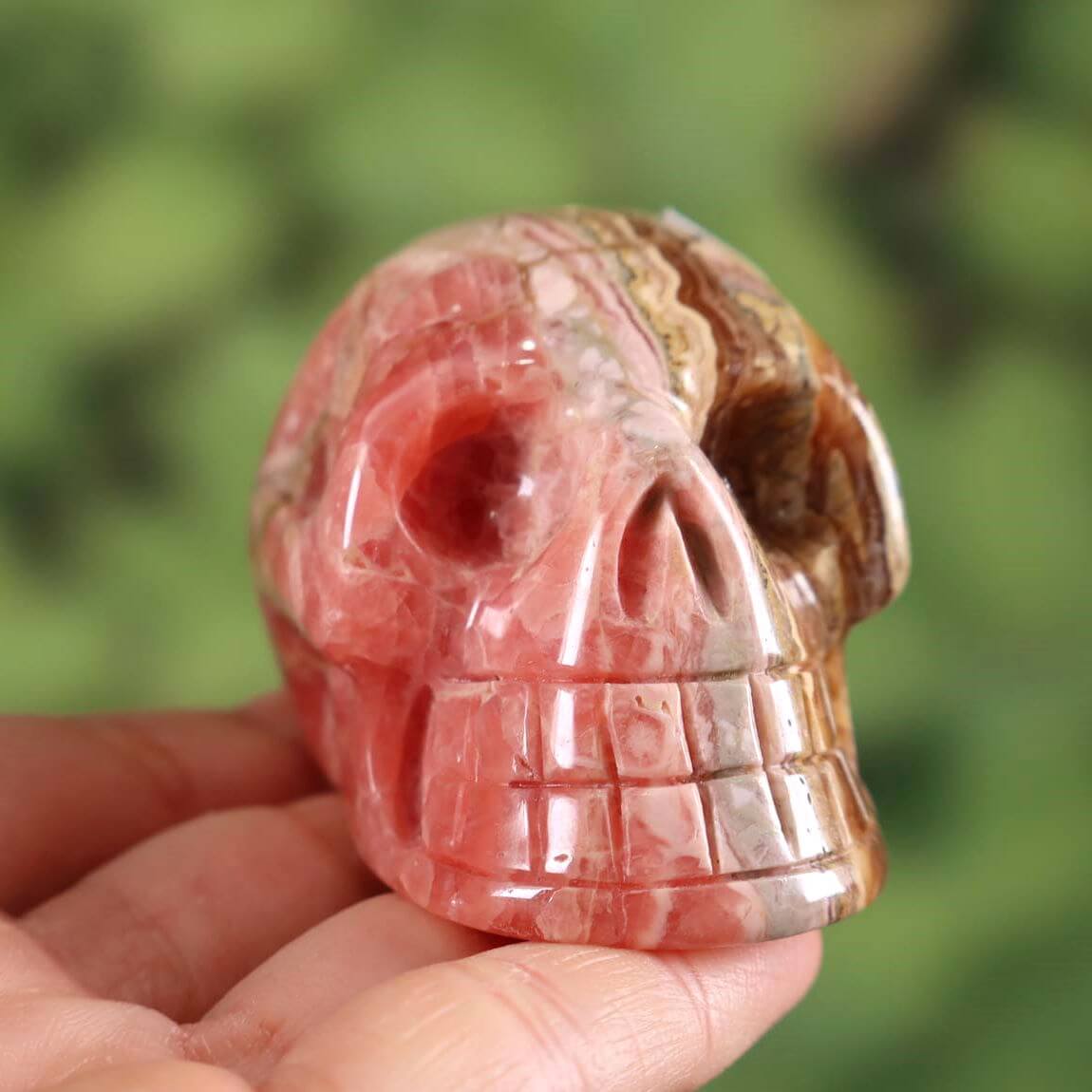 Handcrafted rhodochrosite crystal skull with unique shape, collector's item, 5x4 cm, held in hand against a green background.