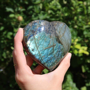 Hand holding large Labradorite heart gemstone displaying vibrant blue and green hues in sunlight.