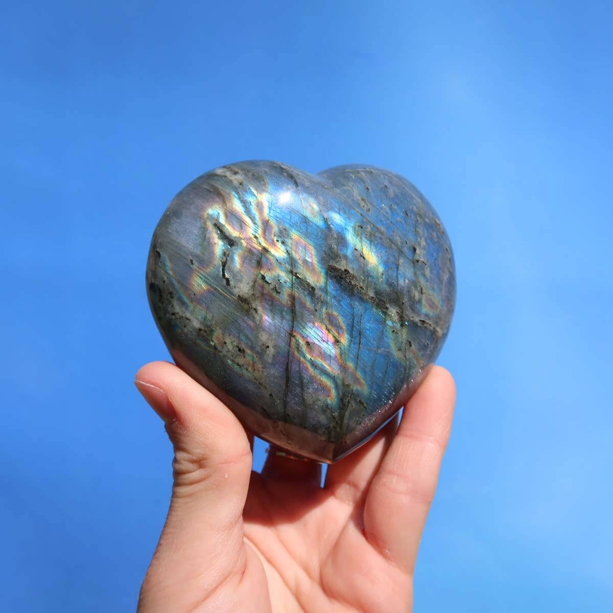 Hand holding a large heart-shaped Labradorite gemstone with unique blue, green, and gold hues on a blue background.