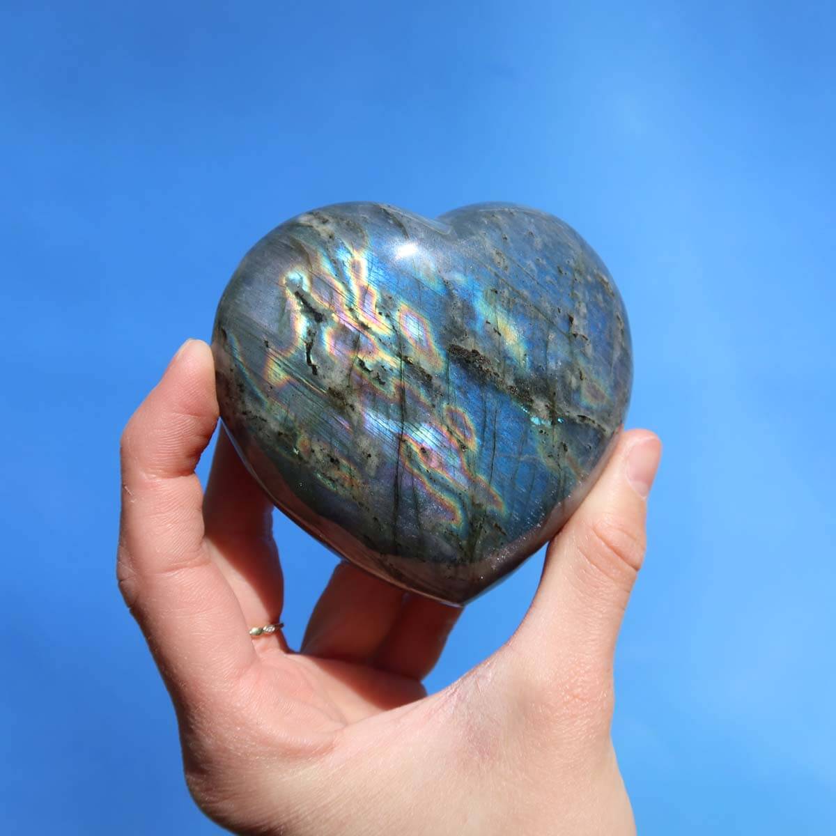 Hand holding a large heart-shaped Labradorite gemstone with unique blue, green, and gold hues on a blue background.