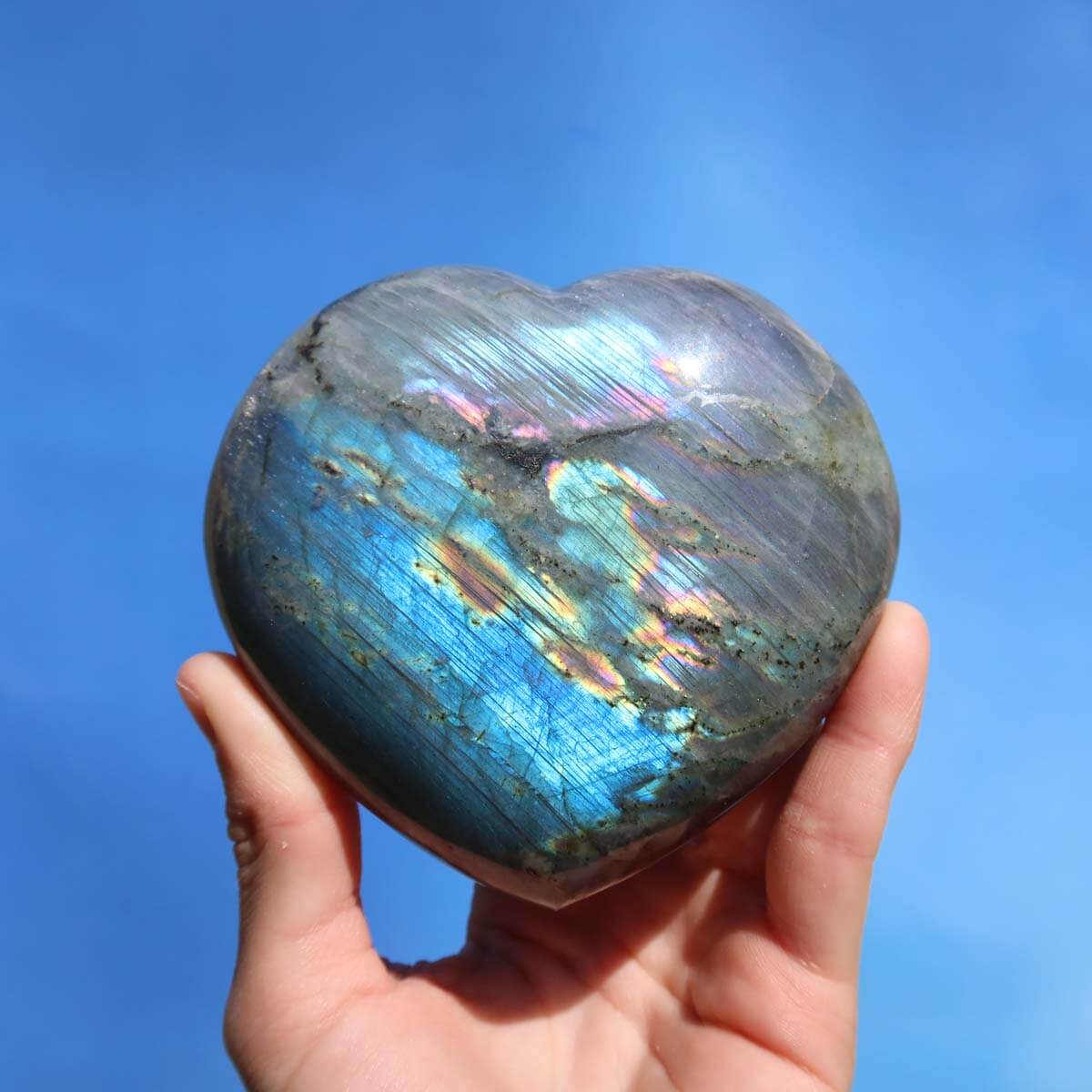 Hand holding a large Labradorite heart gemstone displaying iridescent blue and green colors against a clear sky background.