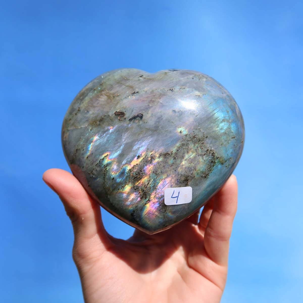 Hand holding a large Labradorite heart gemstone displaying iridescent blue and green colors against a clear sky background.