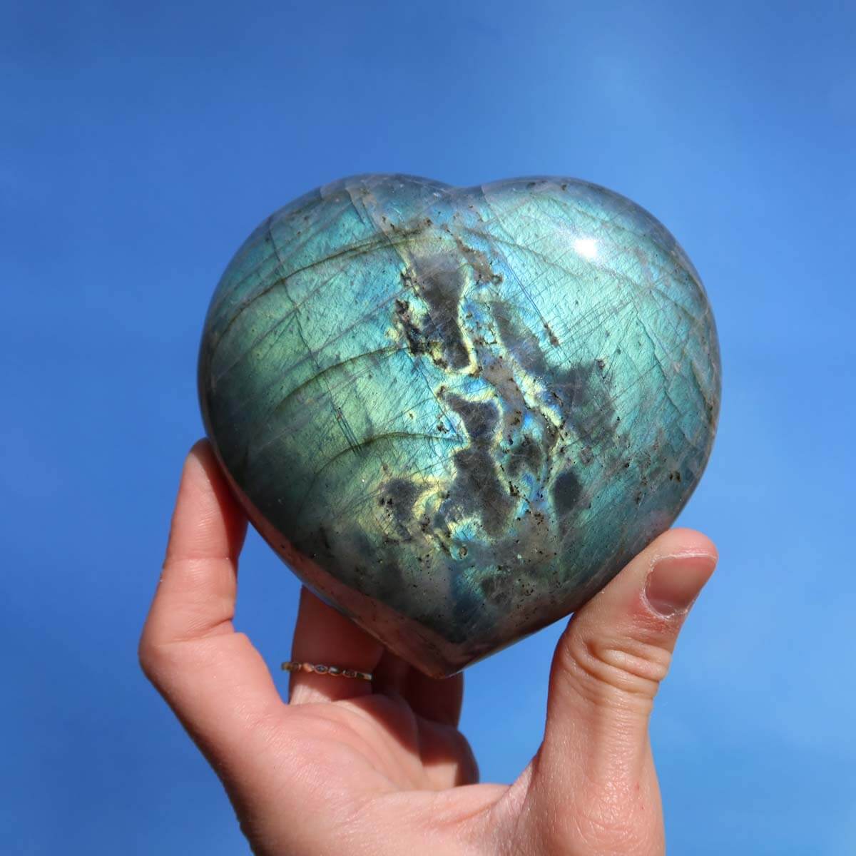 Hand holding large Labradorite heart stone with blue-green hues and unique patterns against a clear blue sky.