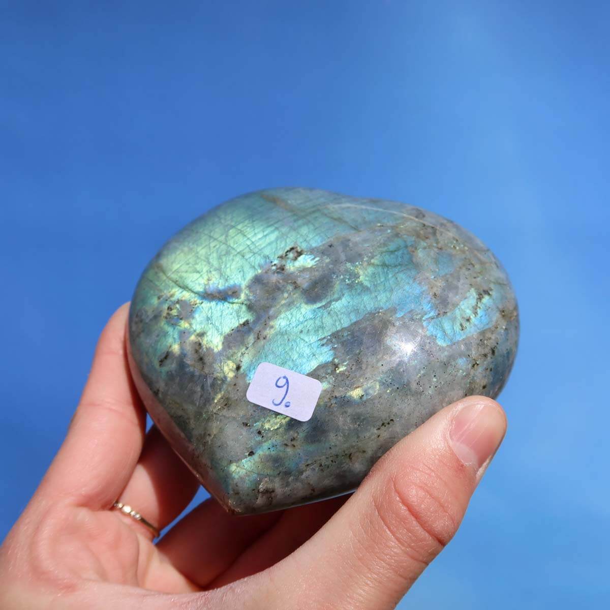 Hand holding large Labradorite heart stone with blue-green hues and unique patterns against a clear blue sky.