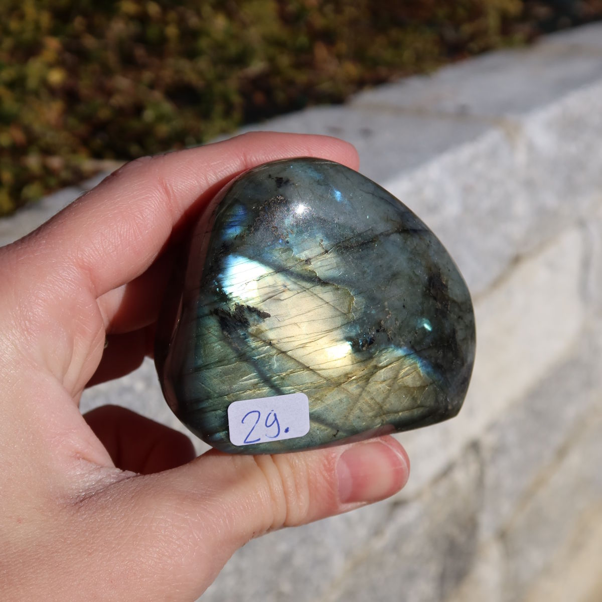 Labradorit Freiform poliert - 5x6cm - Intuition und Kreativität Nr.29 mit schimmernden Farbspielen auf einem Steinuntergrund im Freien.