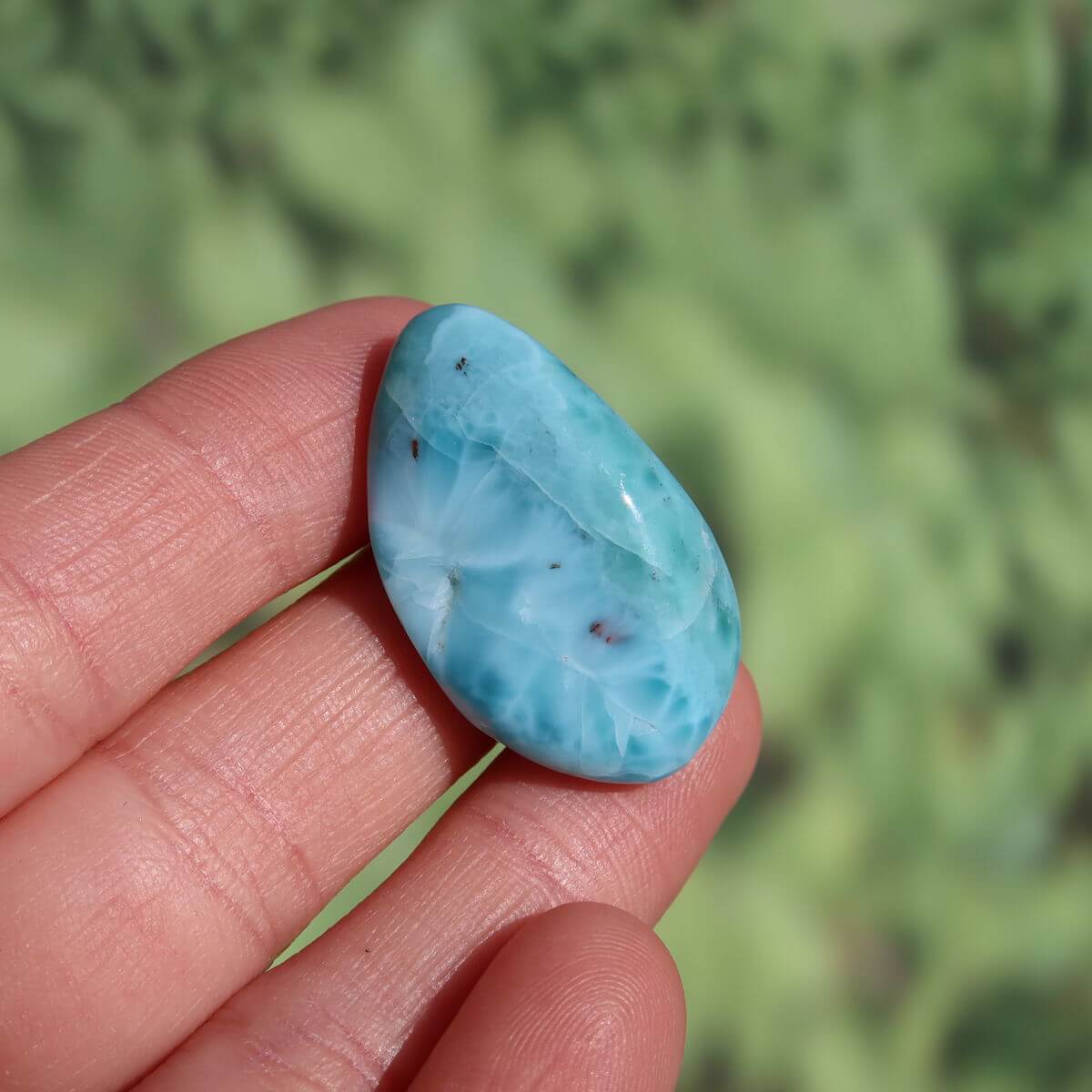Hand holding a 30mm Larimar stone, showcasing its blue hues and unique patterns, against a blurred green background.