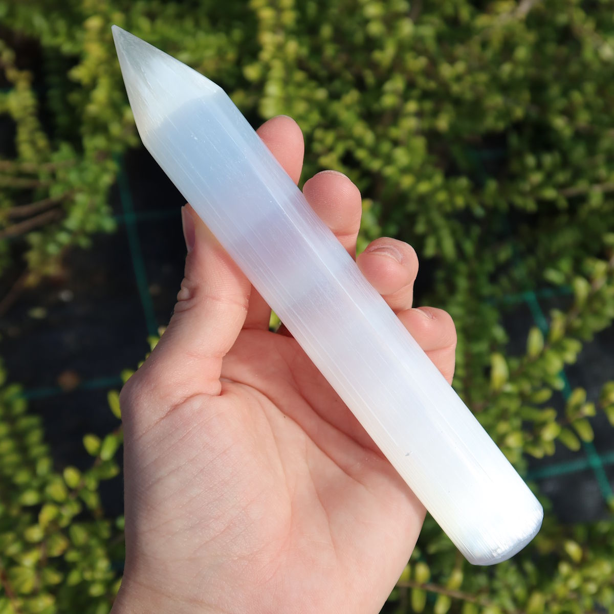 Selenit Massage Griffel mit Spitze- Akupunktur-Massagestab - 15cm in der Hand gehalten vor grünem Hintergrund