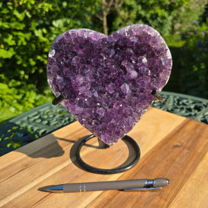 Amethyst Druse Amethyst Geode in Herzform mit Metallsockel 18x16x10cm - NR-43 auf Holzfläche mit Stift im Vordergrund.