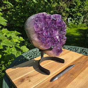 Amethyst Druse Amethyst Geode in Herzform mit Metallsockel 23x19x12cm - NR-18 auf einem Holztisch im Freien.