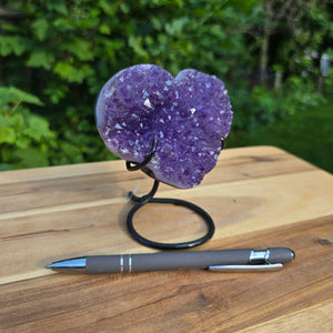Amethyst Druse Amethyst Geode in Herzform mit Metallsockel 12x10x9cm - NR-52 auf Holzoberfläche nebeneinander liegend mit Kugelschreiber.