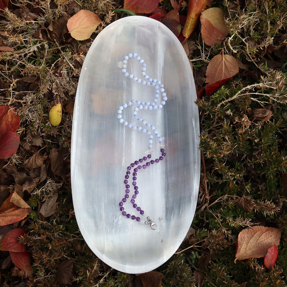 Handgemachte Mala Kette mit Edelsteinen auf einem weißen Tablett, sanfte Farben und klare Linien, Produkt Nr. 23 im Garten gezeigt