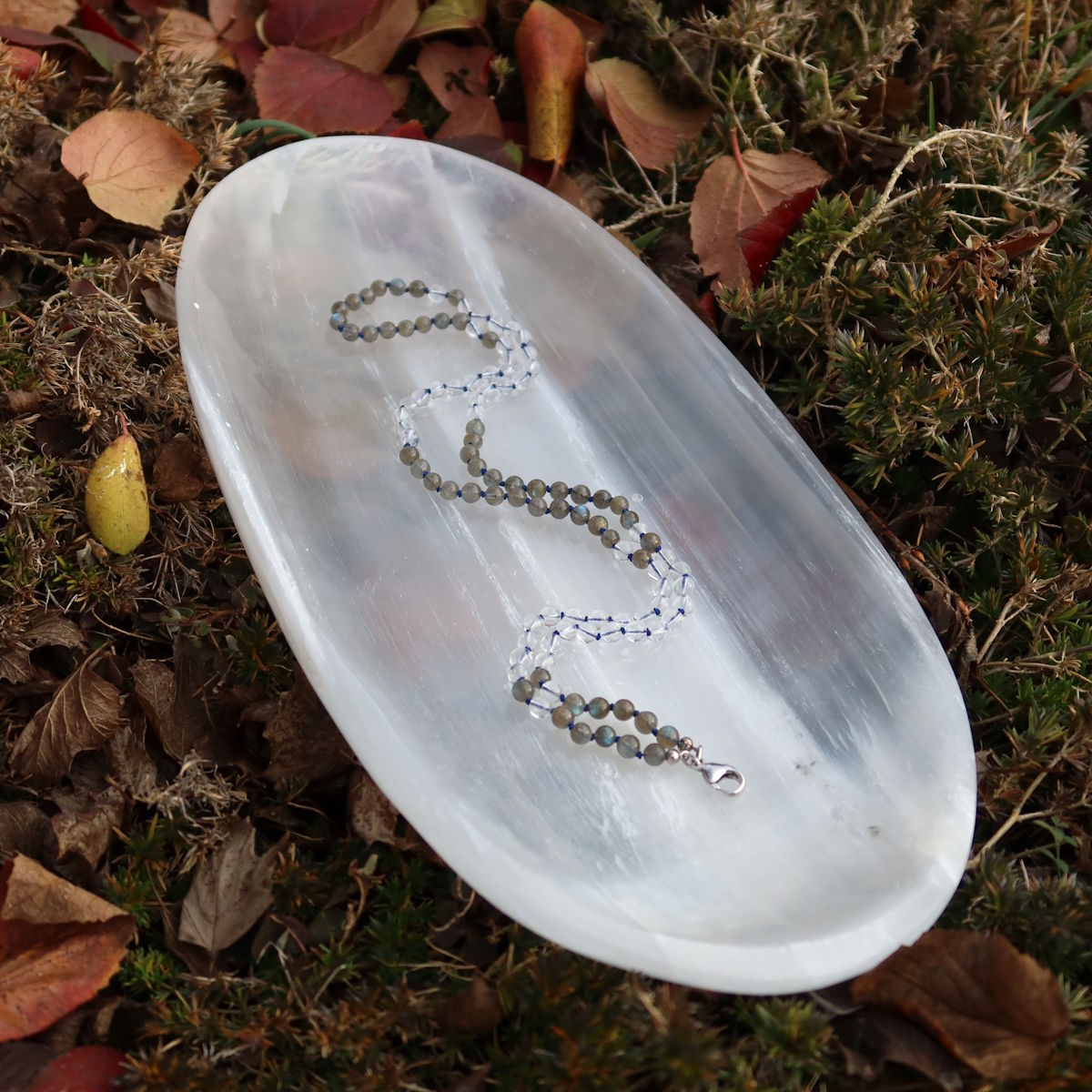 Handgemachte Mala Kette mit Labradorit und Bergkristall auf weißem Teller, umgeben von Blättern, zeigt Kreativität und Klarheit.