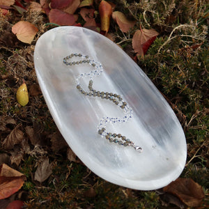 Handgemachte Mala Kette mit Labradorit und Bergkristall auf weißem Teller, umgeben von Blättern, zeigt Kreativität und Klarheit.