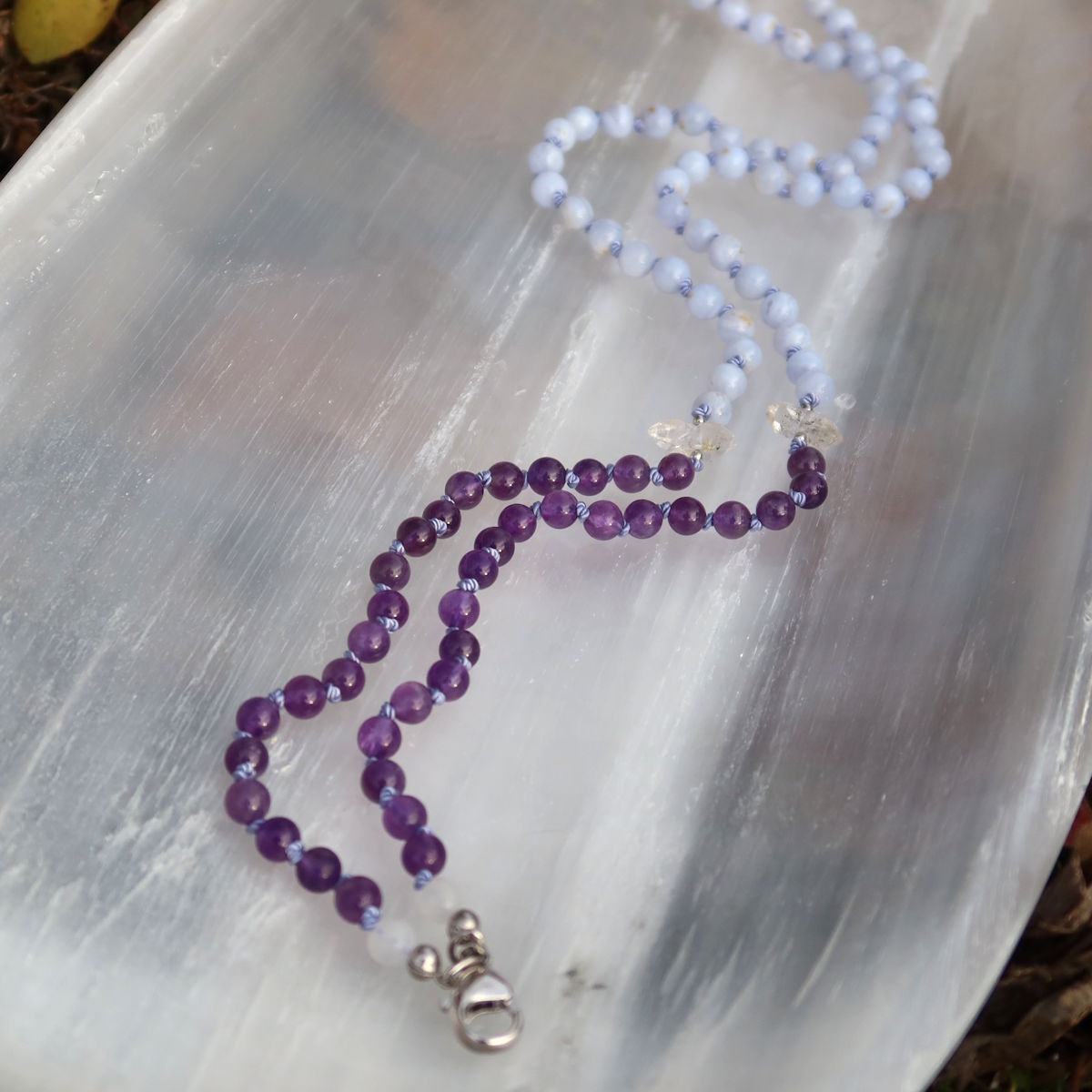 Handgemachte Mala Kette mit Edelsteinen auf einem weißen Tablett, sanfte Farben und klare Linien, Produkt Nr. 23 im Garten gezeigt