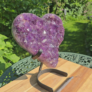 Amethyst Druse Amethyst Geode in Herzform mit Metallsockel 26x23x15cm - NR-22 auf einem Tisch im Garten.