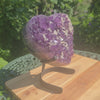 Amethyst Druse Amethyst Geode in Herzform mit Metallsockel 22x20x12cm - NR-6 auf Tisch im Garten.